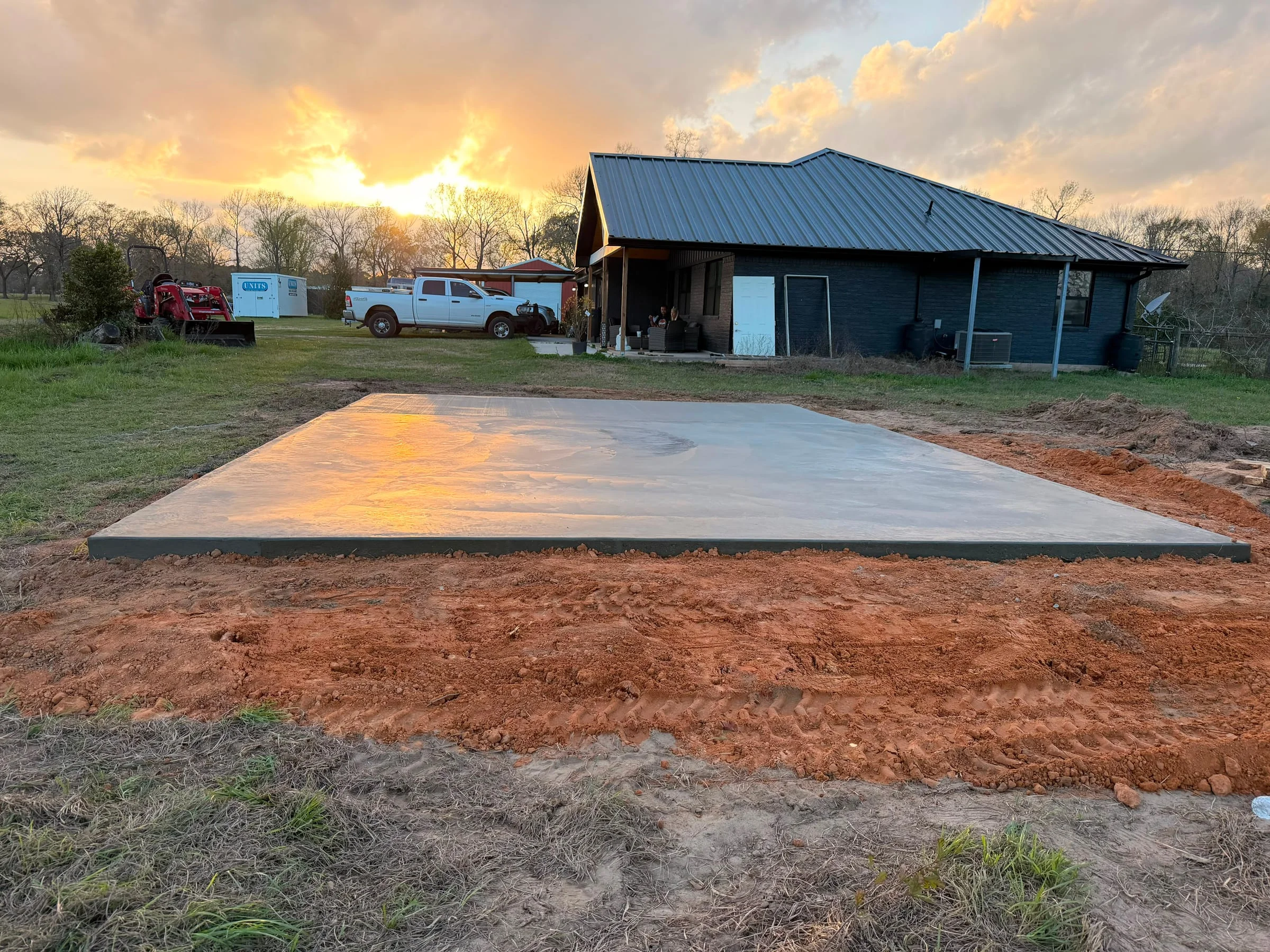 A freshly poured concrete slab on rural acreage at sunset, with the excavated red East Texas clay still banked around its edges